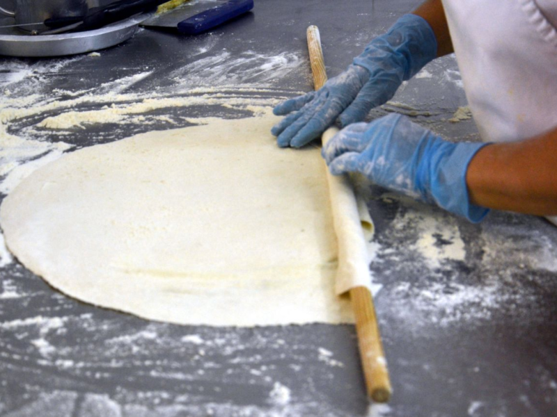 Baker in blue gloves rolling out dough with a wooden rolling pin on a floured surface.